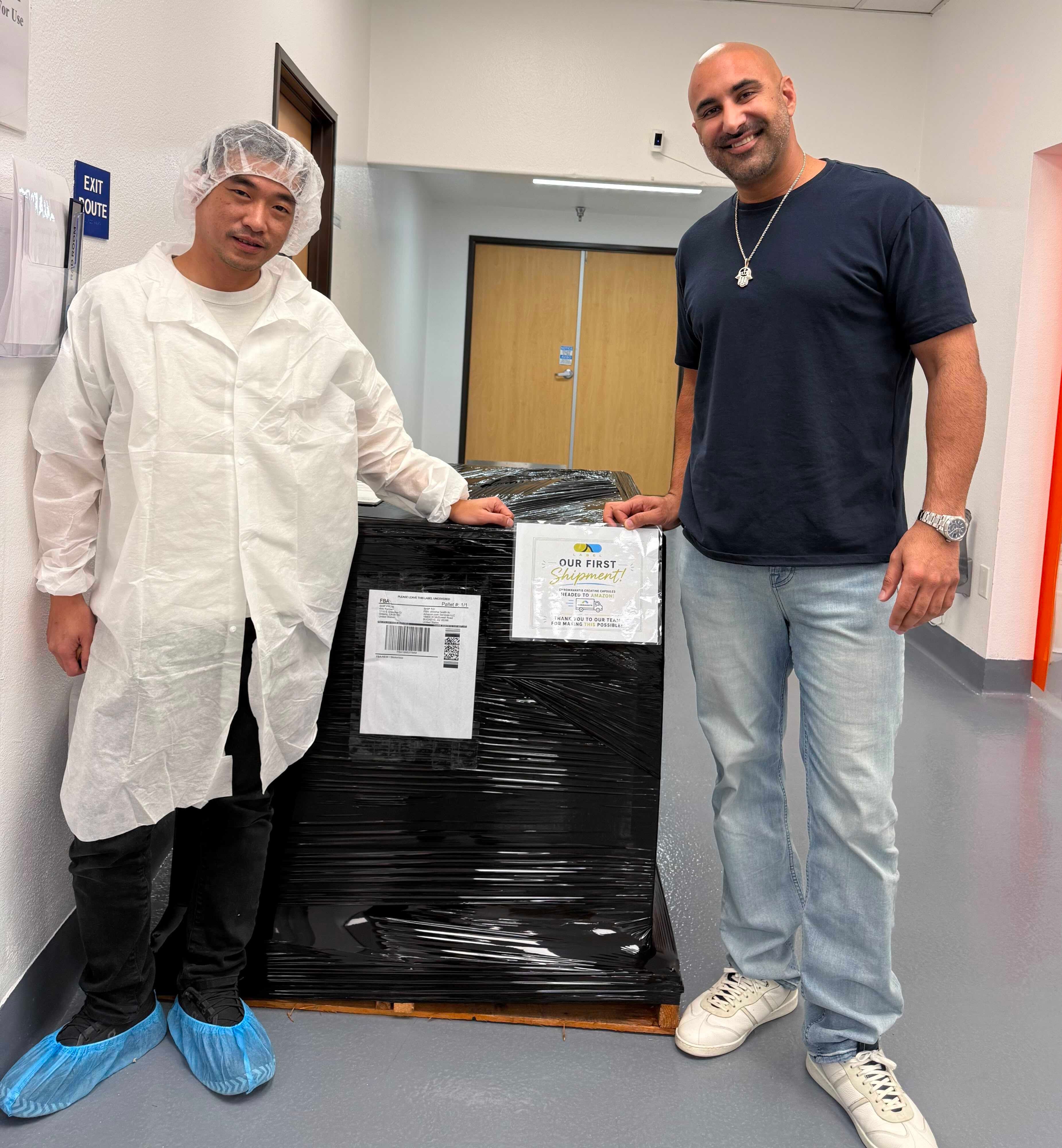 Shah R. standing beside his first shrink-wrapped pallet, labelled for Amazon FBA, with a sign reading Our First Shipment, Headed To Amazon